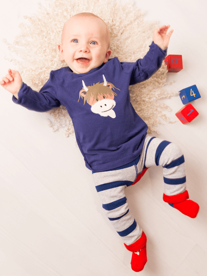Baby wearing a blue shirt with a cow print and striped pants, lying on a white surface with colorful blocks.